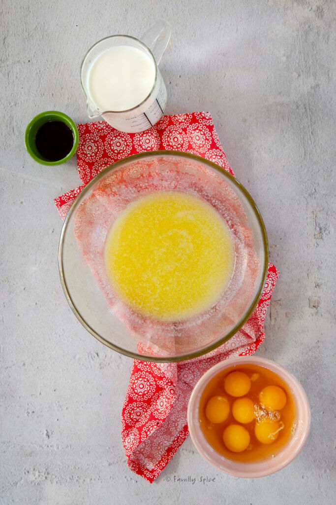 A large glass mixing bowl with melted butter in it with a bowl of raw eggs, a measuring cup with milk and a small bowl of vanilla extract next to ti