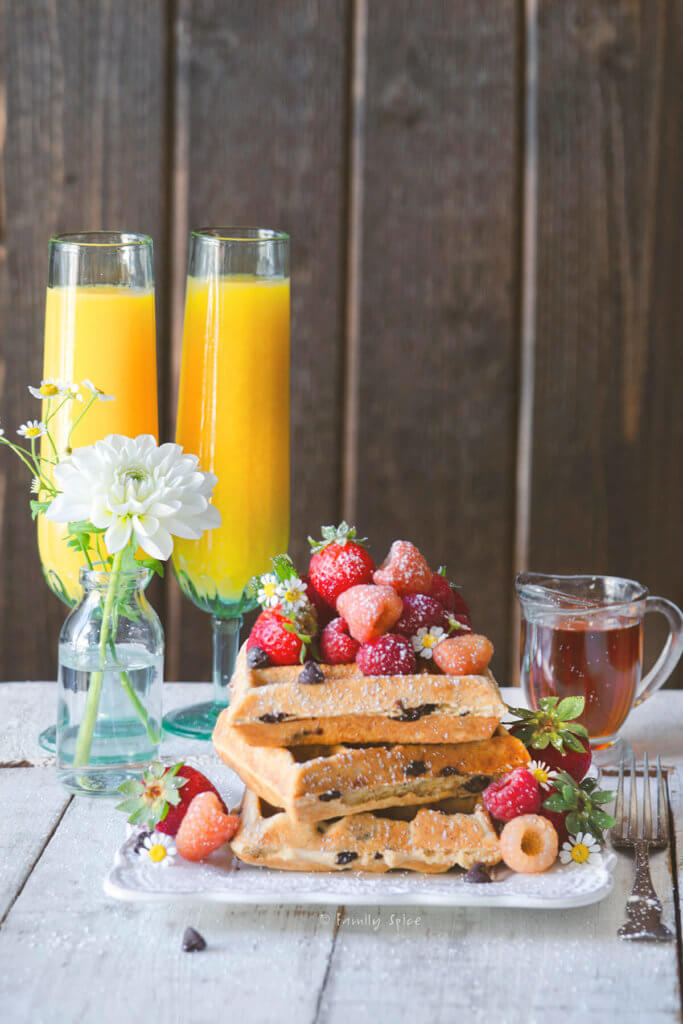 Side view of a stack of chocolate chip waffles topped with fresh berries with flutes of orange juice and a carafe of maple syrup behind it it