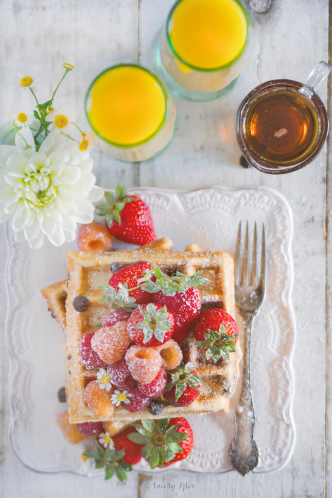Top view of a stack of chocolate chip waffles topped with fresh berries with glasses of orange juice and a carafe of maple syrup next to it