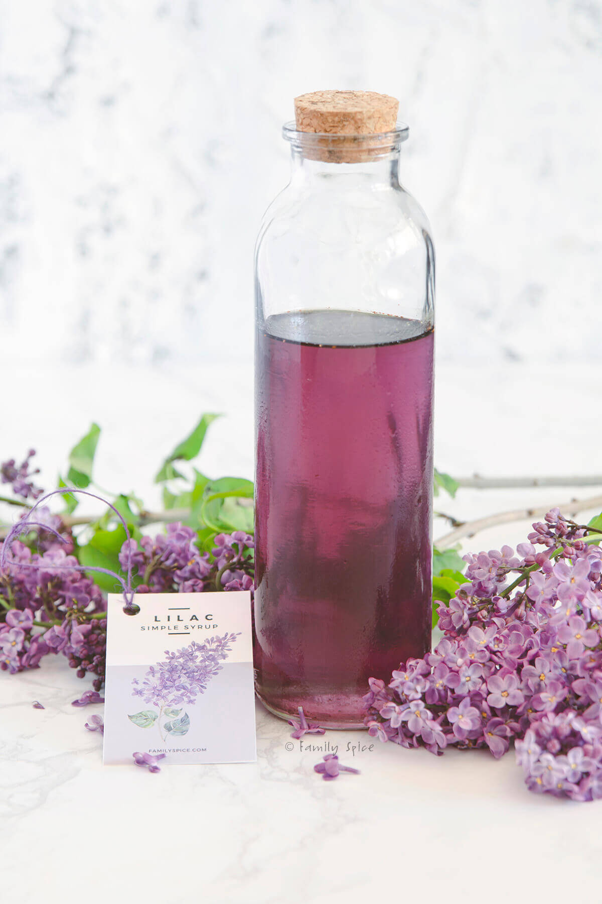 A bottle of lilac syrup with a homemade label and fresh lilacs next to it