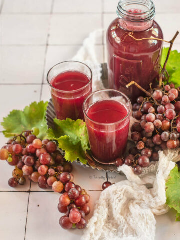 Closeup of a two glasses of homemade grape juice with a bottle of juice on a metal tray with fresh red grapes