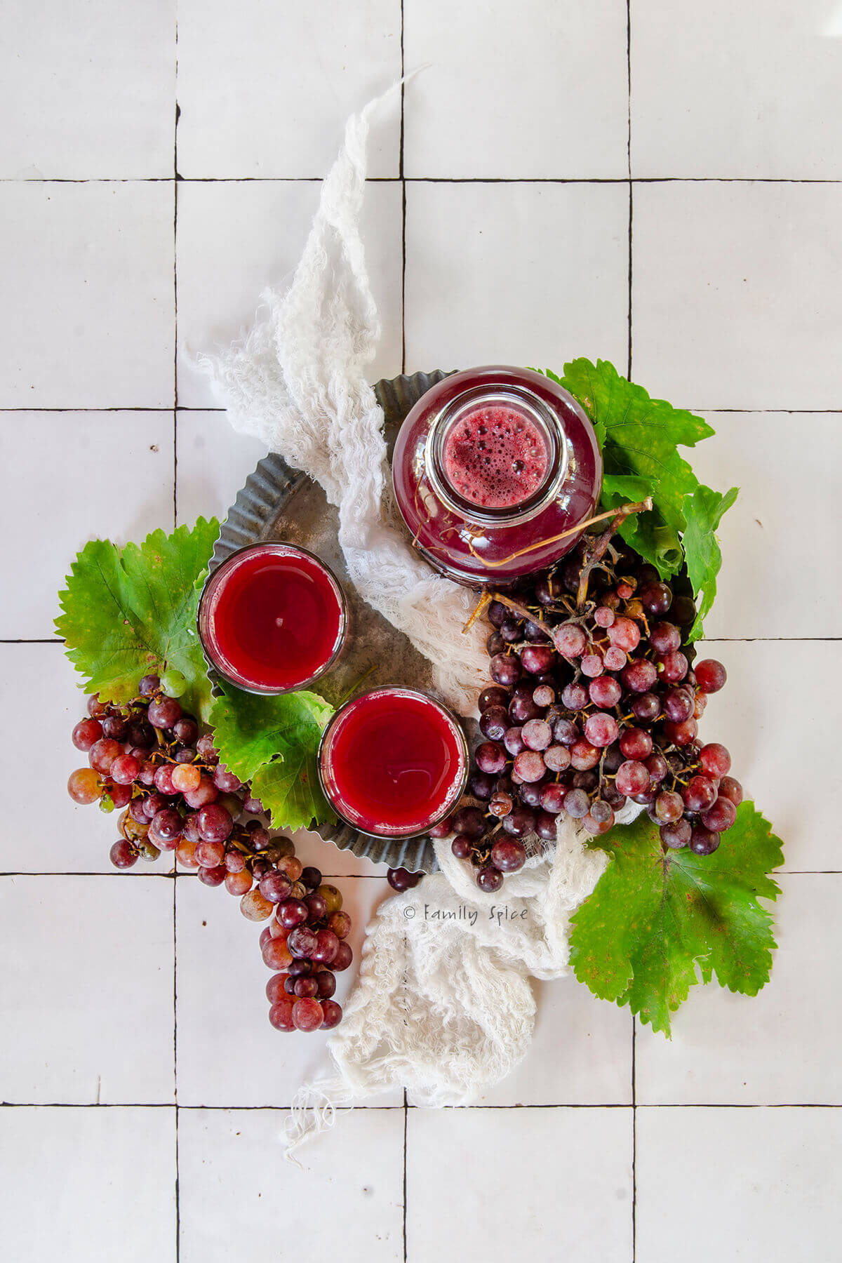 Top view of a bottle of homemade grape juice with two glass of juice on a metal tray with fresh red grapes