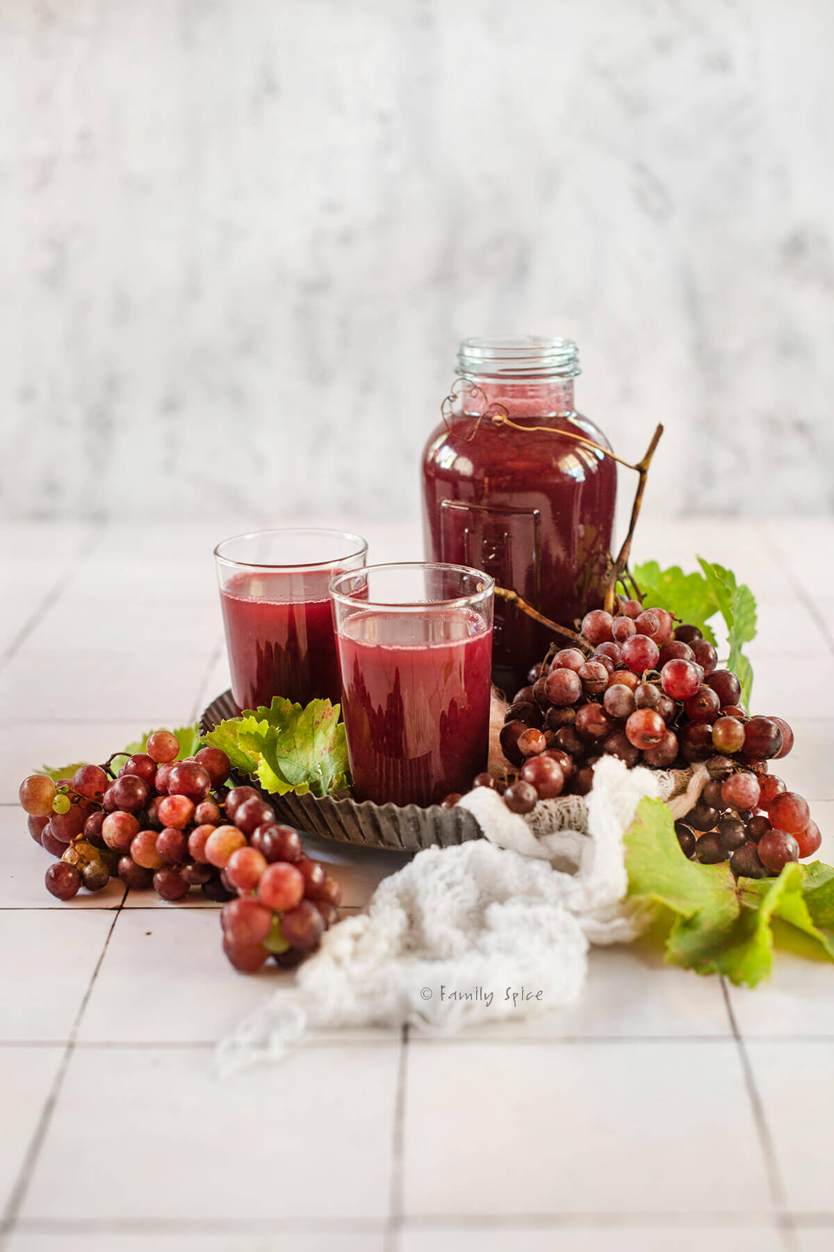 Side view of a bottle of homemade grape juice with two glass of juice on a metal tray with fresh red grapes