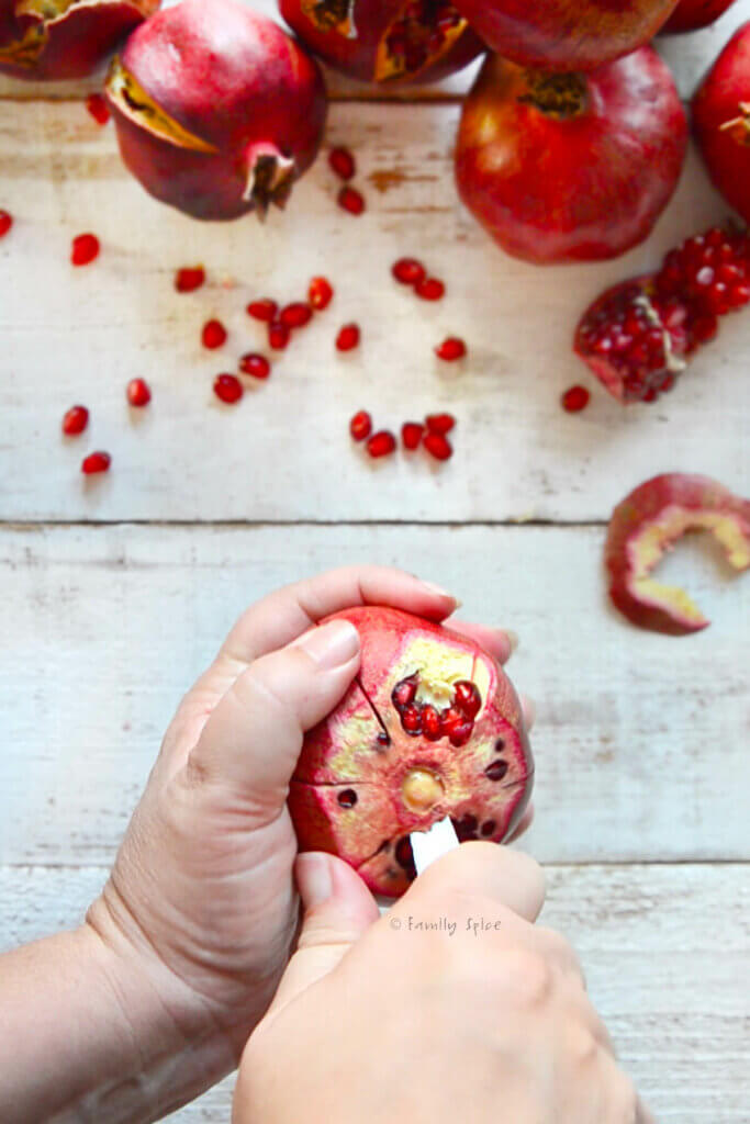 A hand holding a pomegranate with the crown remove and sides scored and using a knife to cut the center out