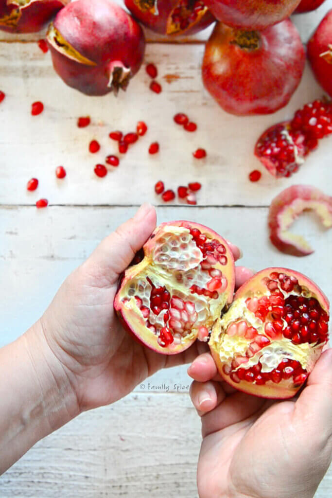 A hand holding a halved pomegranate with more pomegranates next to it