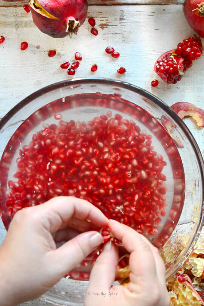 A hand pulling pomegranate arils over a bowl of water with more pomegranate in it