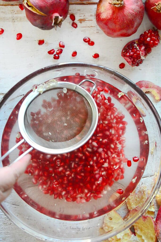 Using a small mesh strainer to strain out pith from a bowl of water with pomegranate arils in it