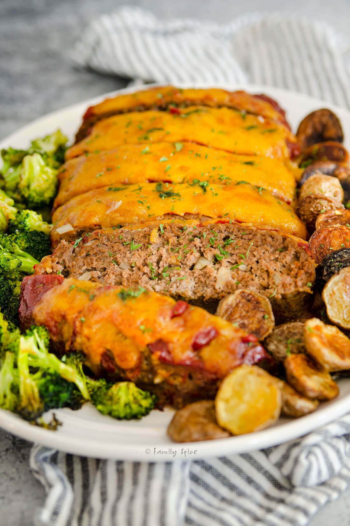 Closeup of a cheeseburger meatloaf cut into slices with roasted potatoes and broccoli next to it