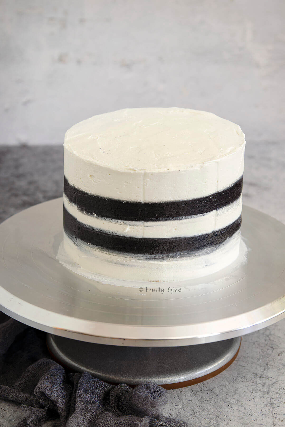 A white frosted cake with 2 black stripes on a stainless cake stand