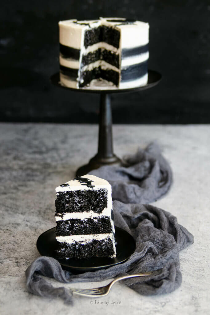 A slice of a 3-layer black and white cake on a black plate with the rest of the cake blurred behind it