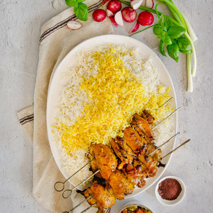 Top view of an oval platter with basmati rice topped with saffron and skewers of chicken kebab on it and fresh herbs and radishes next to it