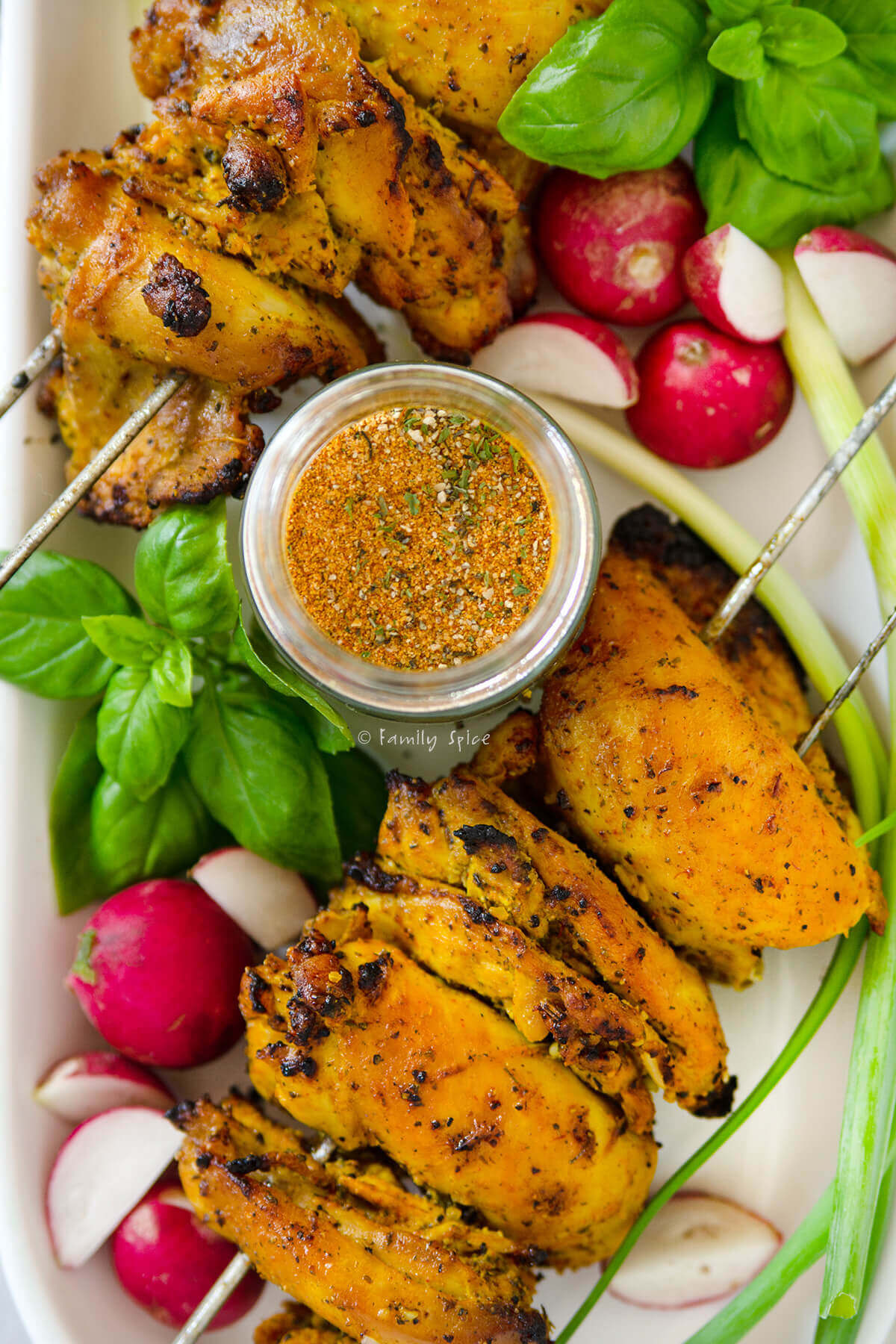 Closeup of a jar of dry rub chicken seasoning on a plate of chicken kebabs and fresh herbs