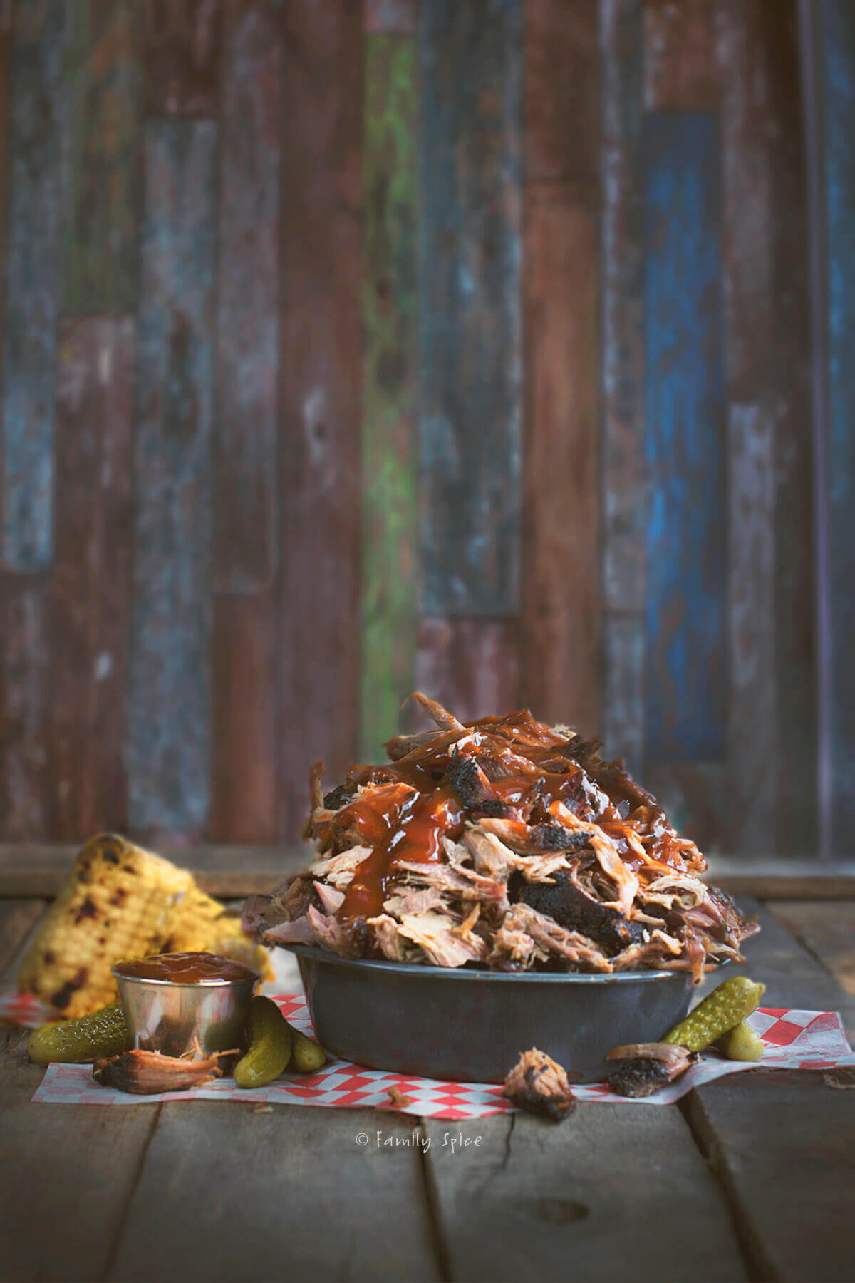 Side view of a metal bowl piled with smoked pulled pork topped with bbq sauce with corn and pickles next to it