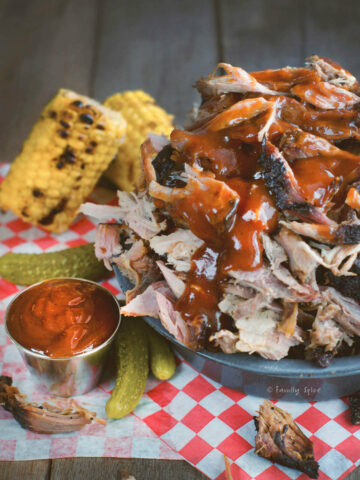 Closeup of a metal bowl piled with smoked pulled pork topped with bbq sauce with corn and pickles next to it