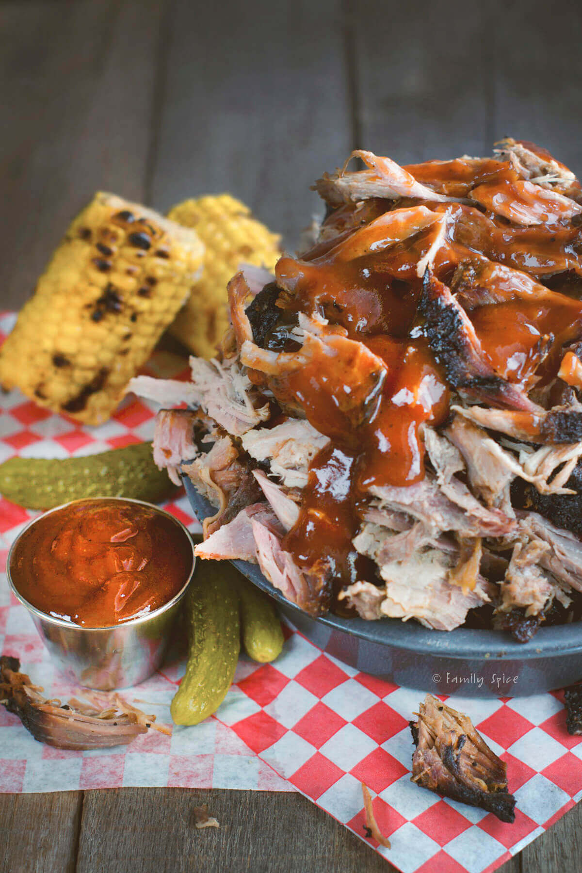Closeup of a metal bowl piled with smoked pulled pork topped with bbq sauce with corn and pickles next to it