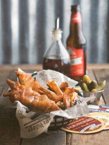 Side view of a basket with beer battered fish and chips in it and beer, malt vinegar and pickles behind it