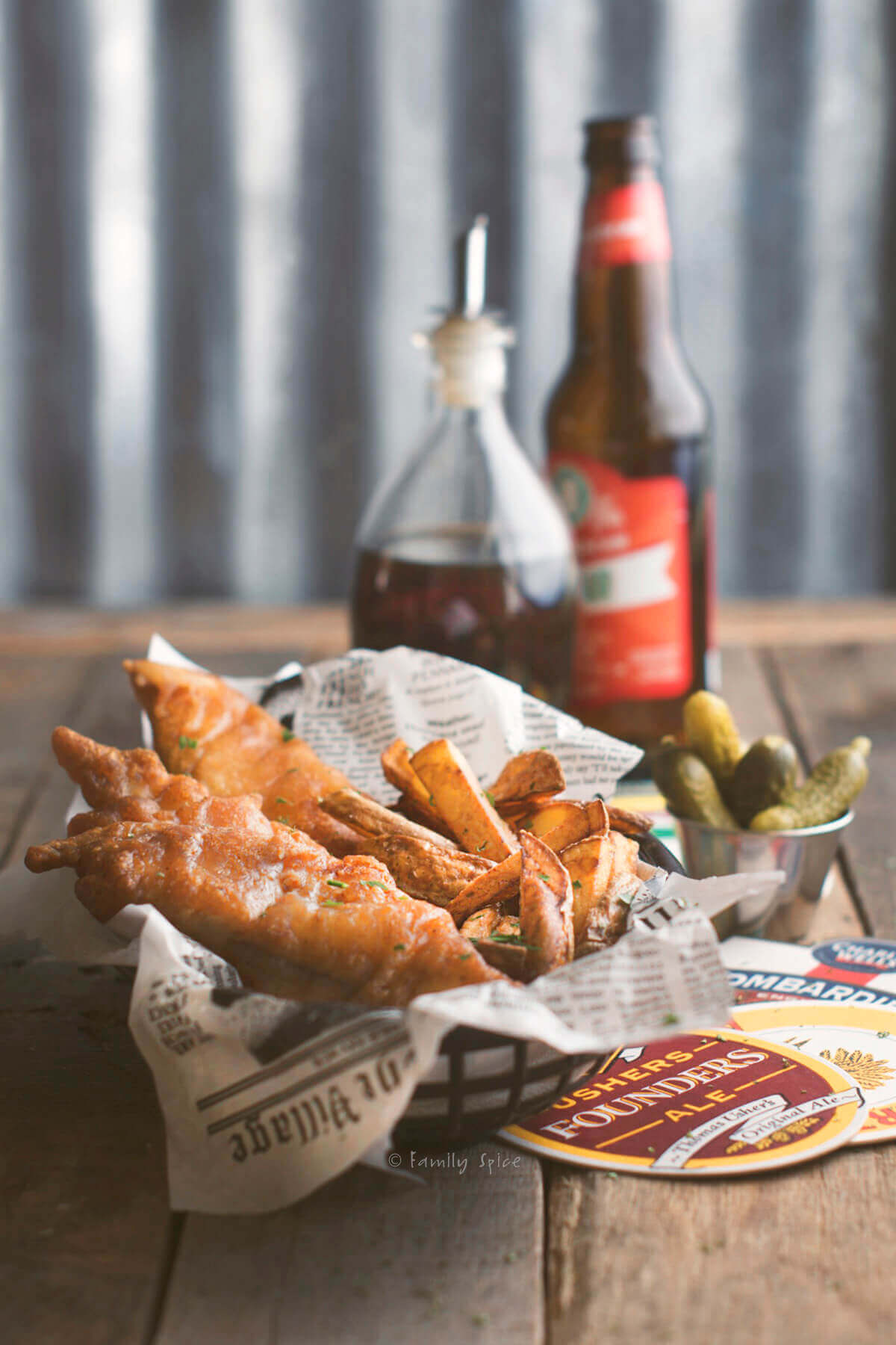 Side view of a basket with beer battered fish and chips in it and beer, malt vinegar and pickles behind it