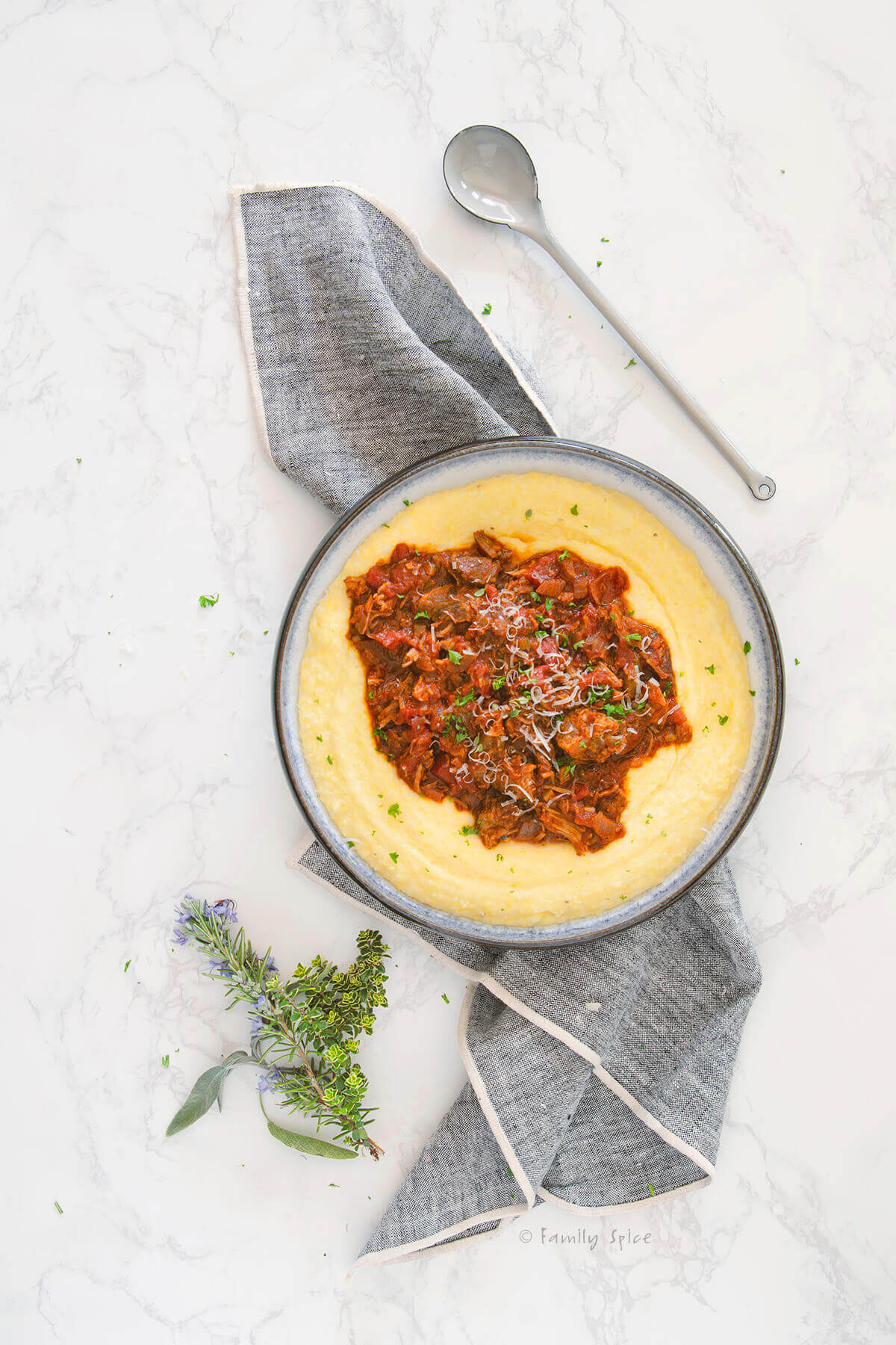 Top view of a shallow bowl with creamy polenta and topped with pork ragu on a napkin with herbs next to it