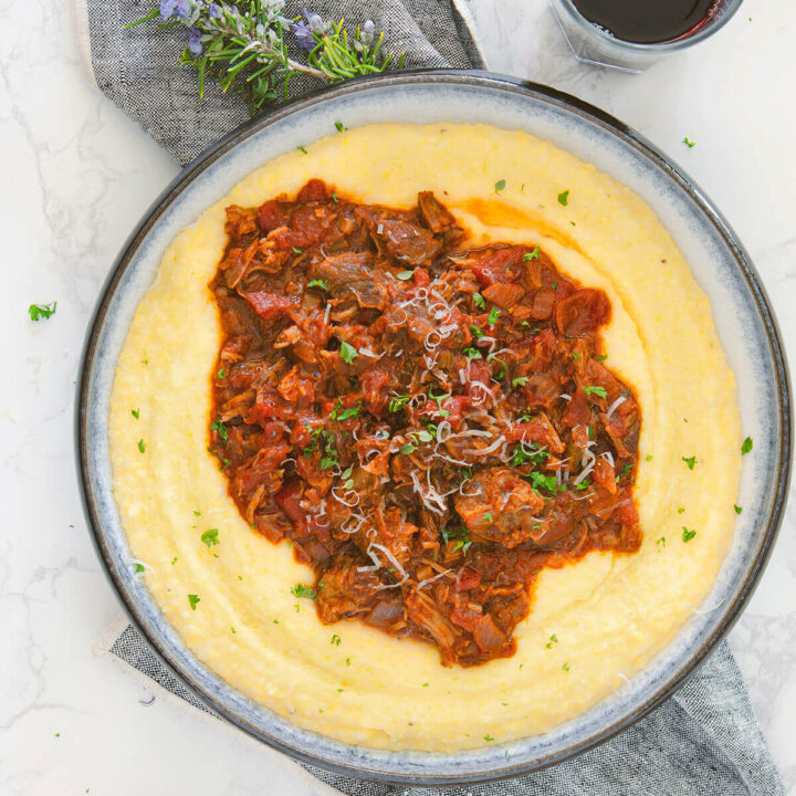 Top view closeup of a shallow bowl with creamy polenta and topped with pork ragu on a napkin with a glass of red wine next to it