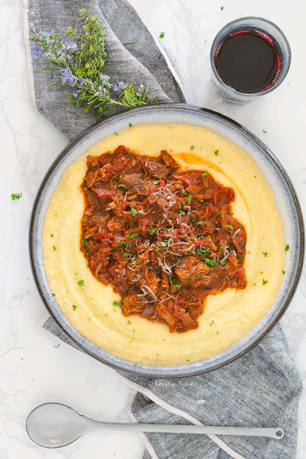 Top view closeup of a shallow bowl with creamy polenta and topped with pork ragu on a napkin with a glass of red wine next to it