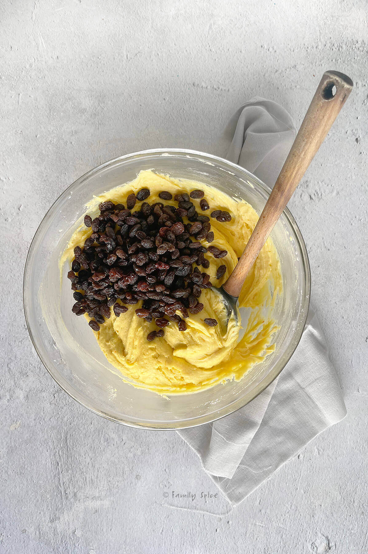A mixing bowl with mixed saffron cake batter and raisins in it and a rubber scraper