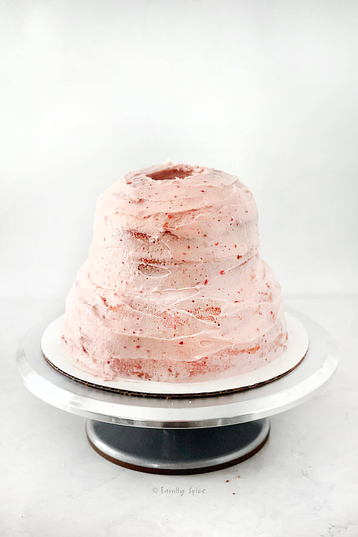 A strawberry cake in a bell shape with the center hollowed out and lightly frosted with strawberry buttercream frosting on a metal cake stand
