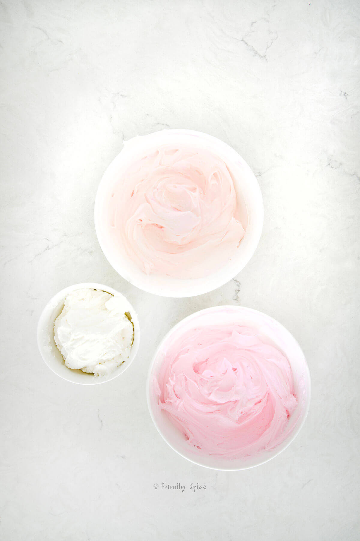 A large bowl of dark pink frosting, a bowl of light pink frosting and a smaller bowl of white frosting