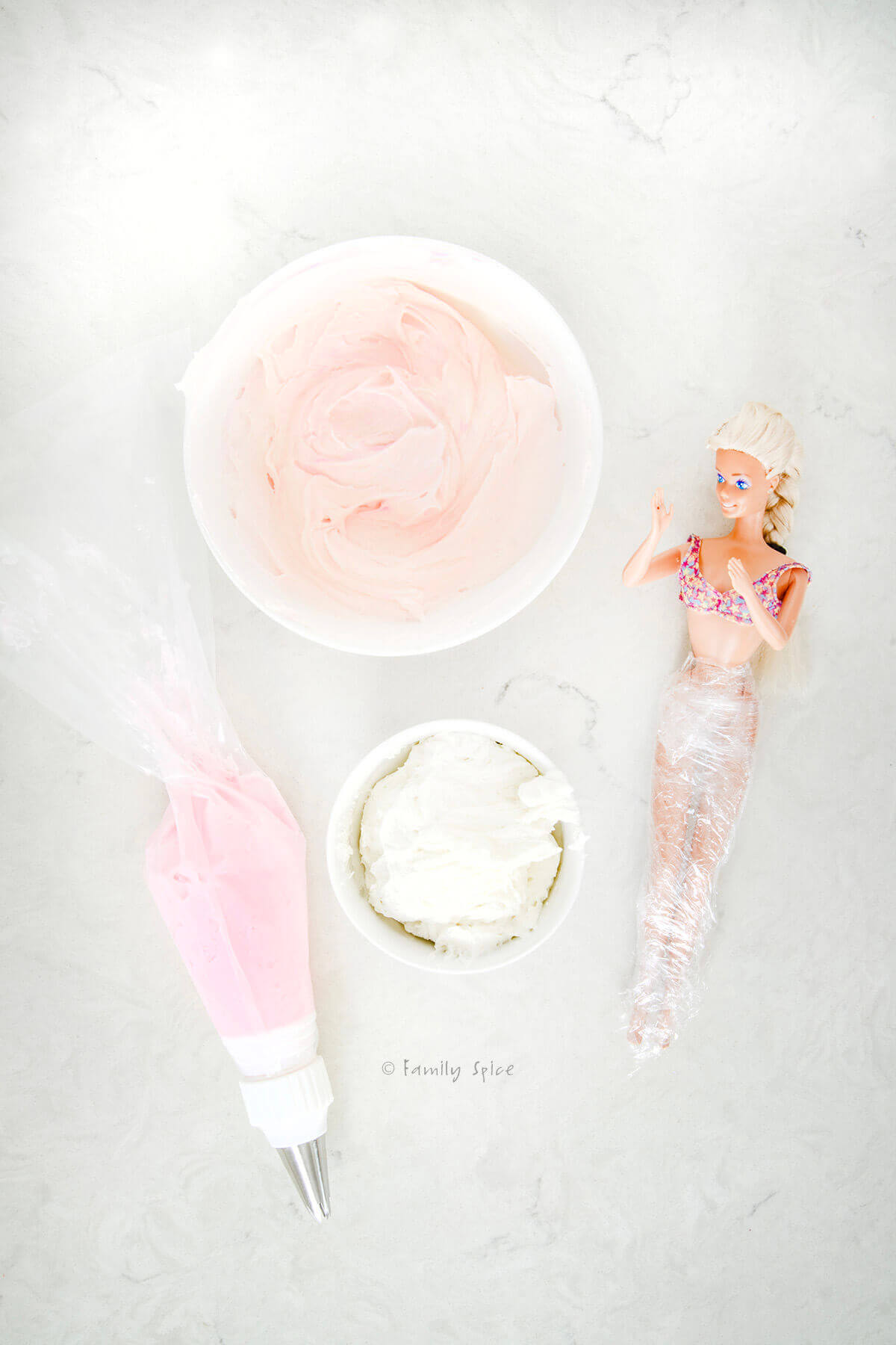 A bowl of light pink frosting, a smaller bowl of white frosting, a piping bag with dark pink buttercream frosting and a barbie with legs wrapped in plastic wrap