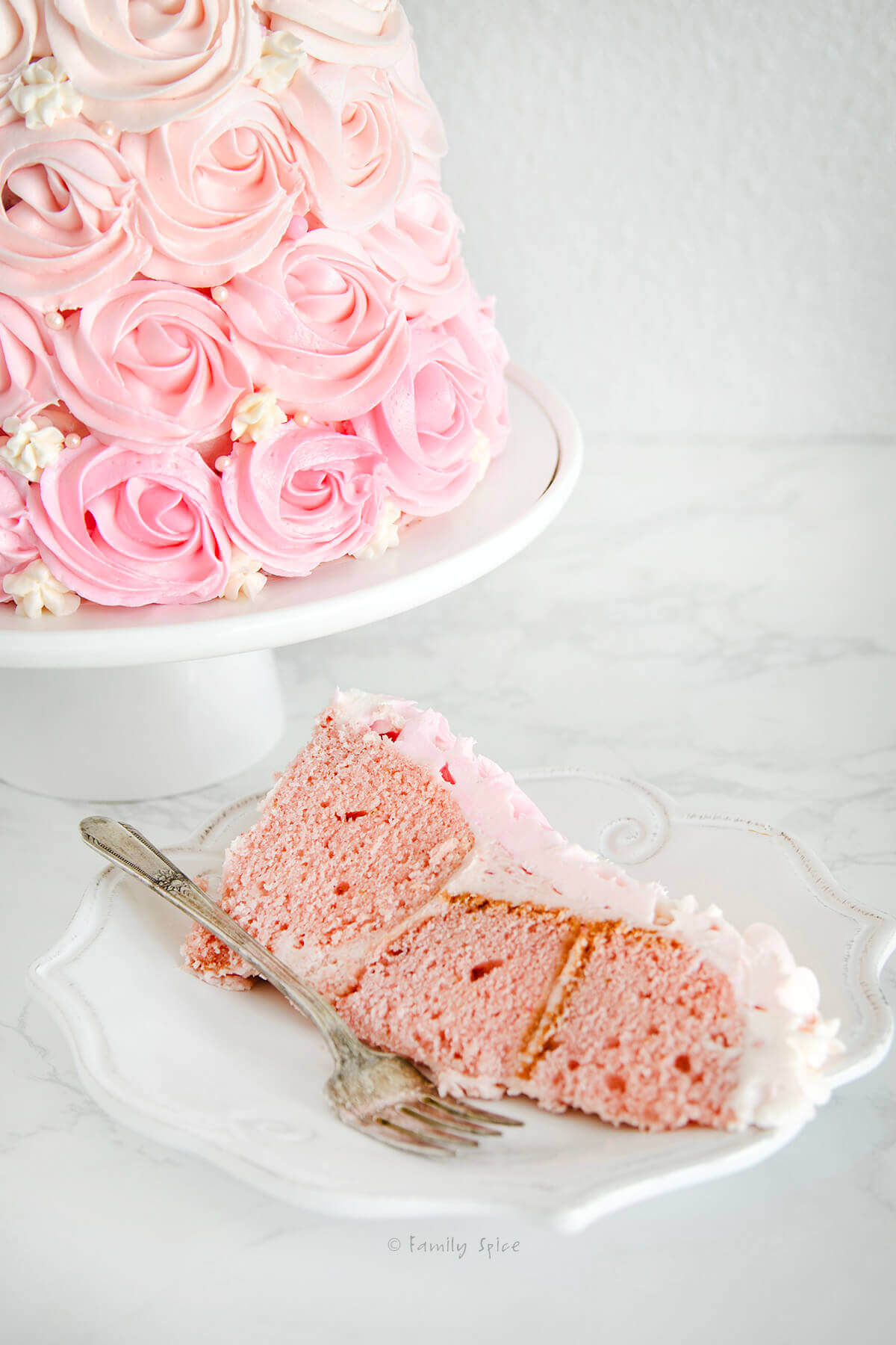 Closeup of a slice of pink strawberry cake o a white plate with the rest of a rose ombre cake on a white cake stand behind it