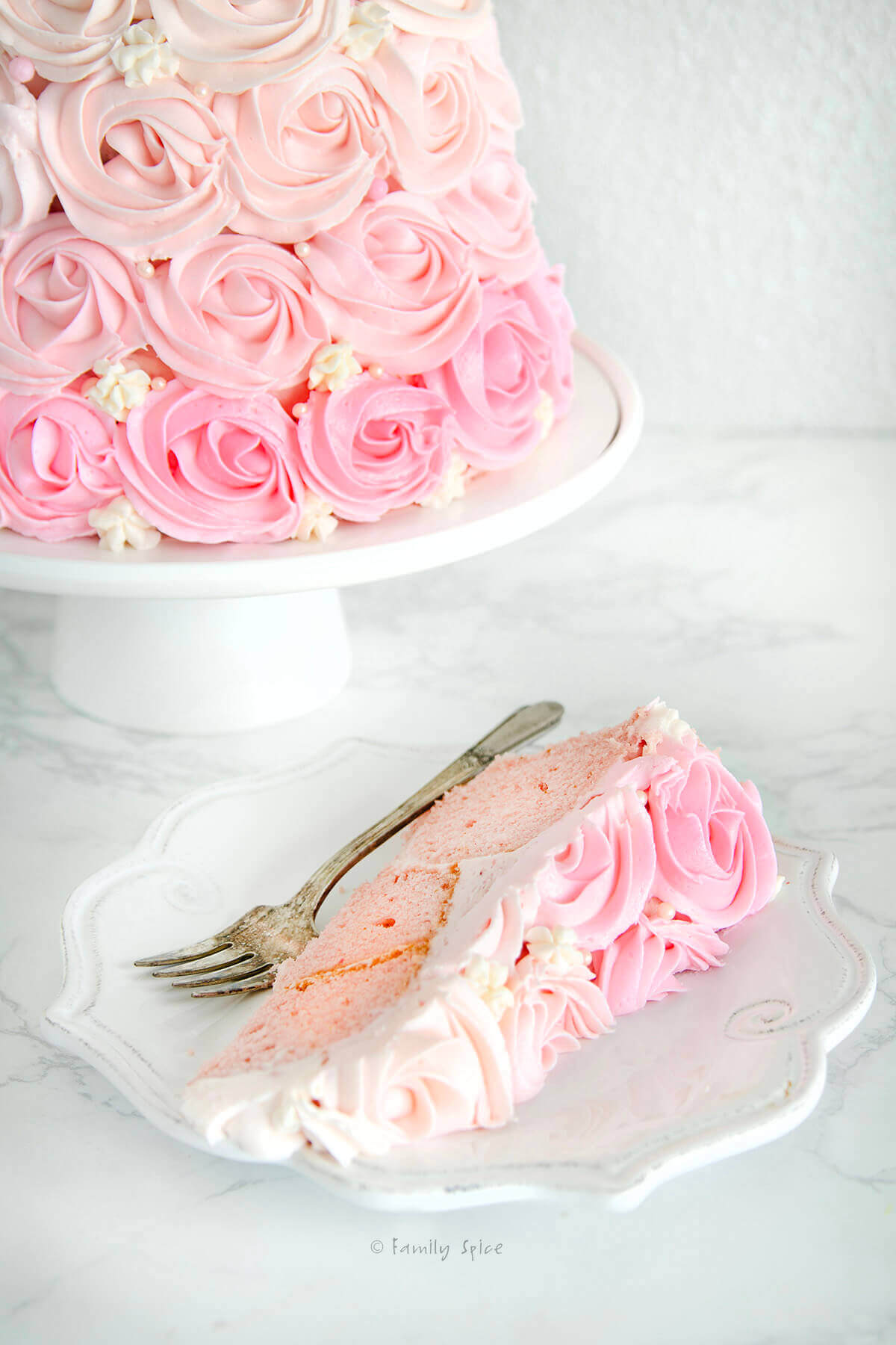 Closeup of a slice of pink ombre cake with the rest of a rose ombre cake on a white cake stand behind it