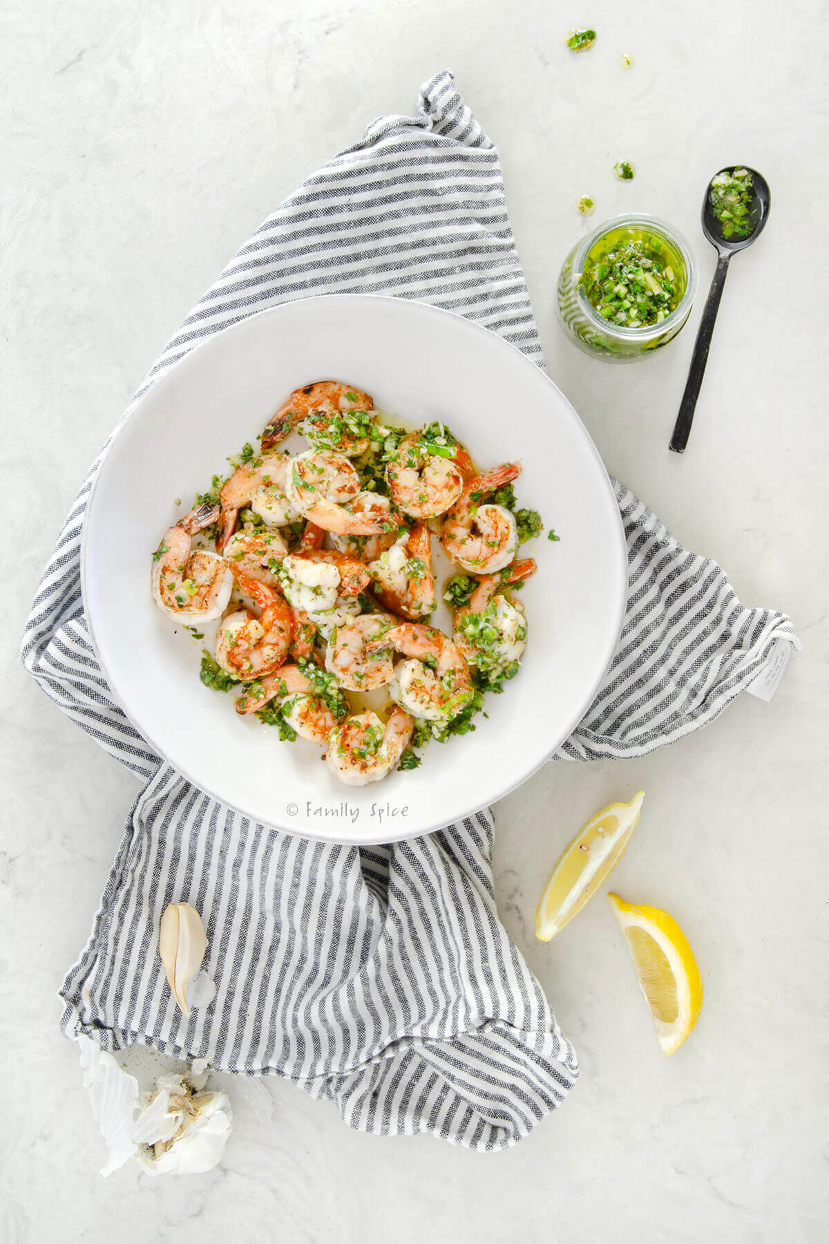 A shallow white bowl with grilled shrimp mixed with cilantro chimichurri in it along with lemon wedges, some garlic and small jar of chimichurri in it