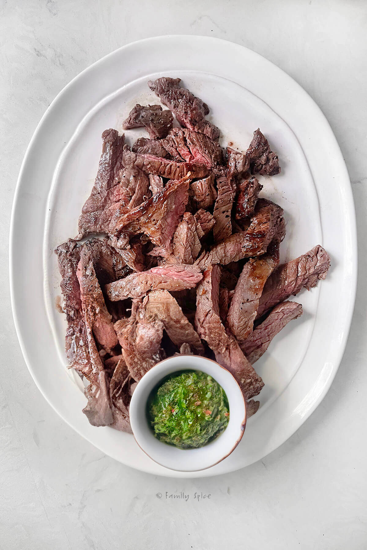 An oval white platter with slices of cooked flank steak with a small bowl of cilantro chimichurri with it
