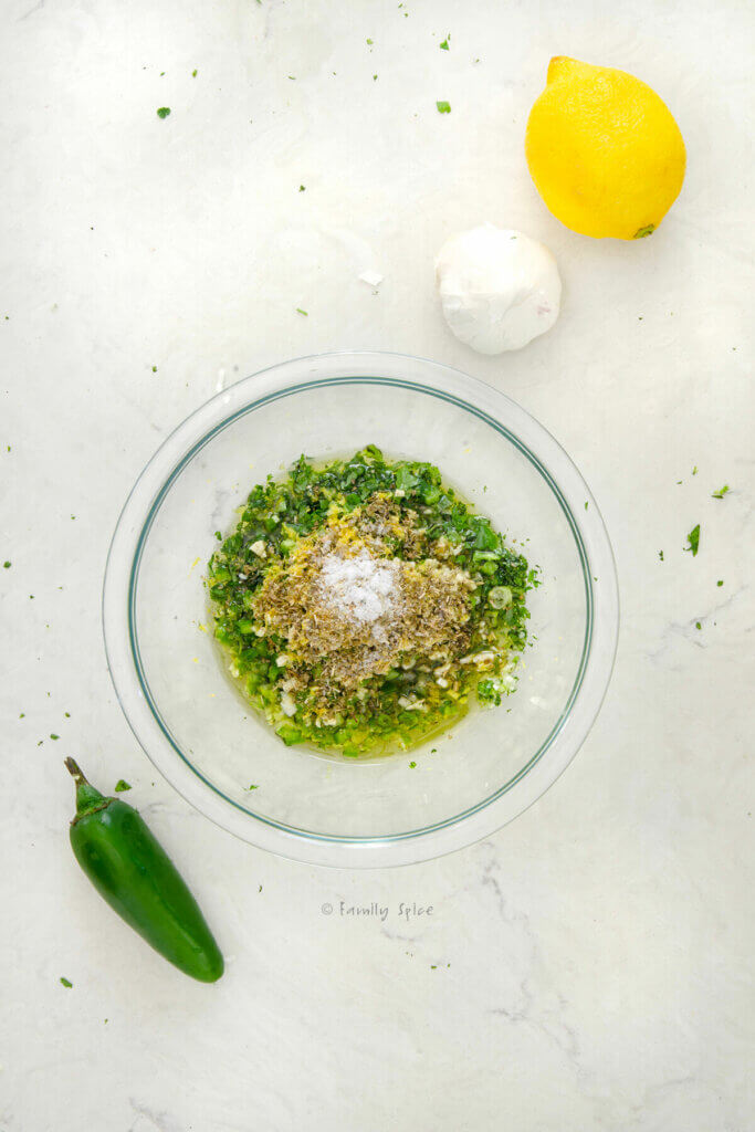 Top view of a small glass bowl with ingredients to make cilantro chimichurri mixed in it and with a lemon, jalapeño and garlic next to it