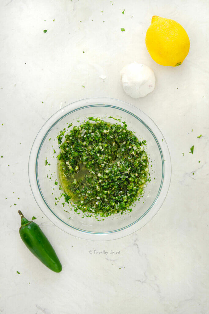 Top view of a small glass bowl with cilantro chimichurri mixed in it and with a lemon, jalapeño and garlic next to it