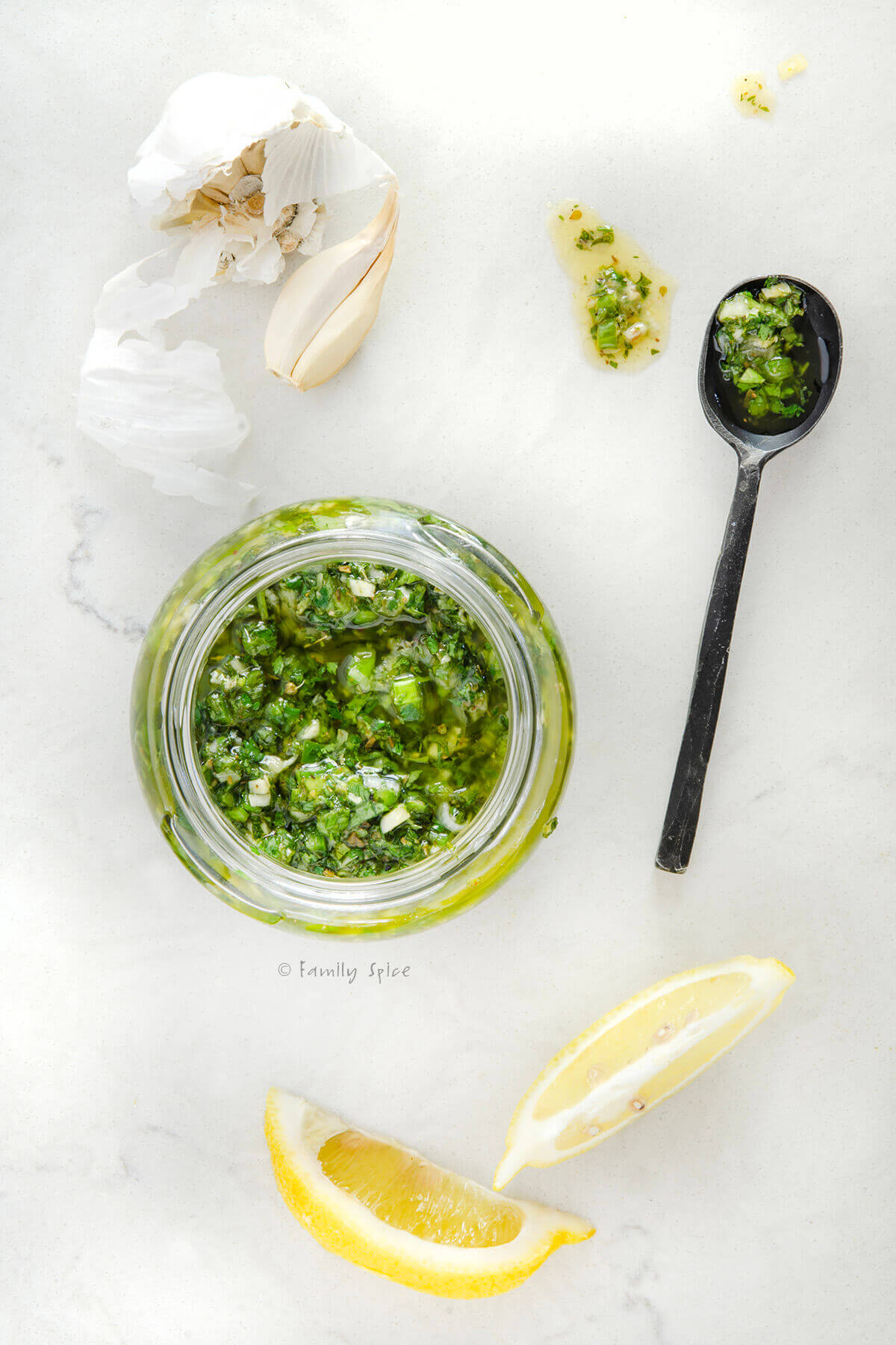 Top view of a jar of cilantro chimichurri with a metal spoon, lemon wedges and garlic next to it