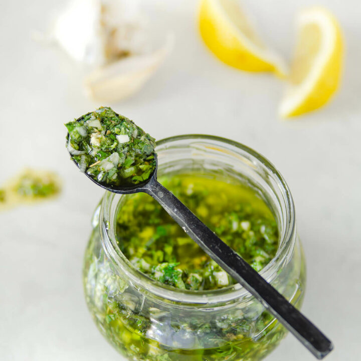 Closeup of a jar of cilantro chimichurri with a metal spoon on top of it and lemon wedges and garlic next to it
