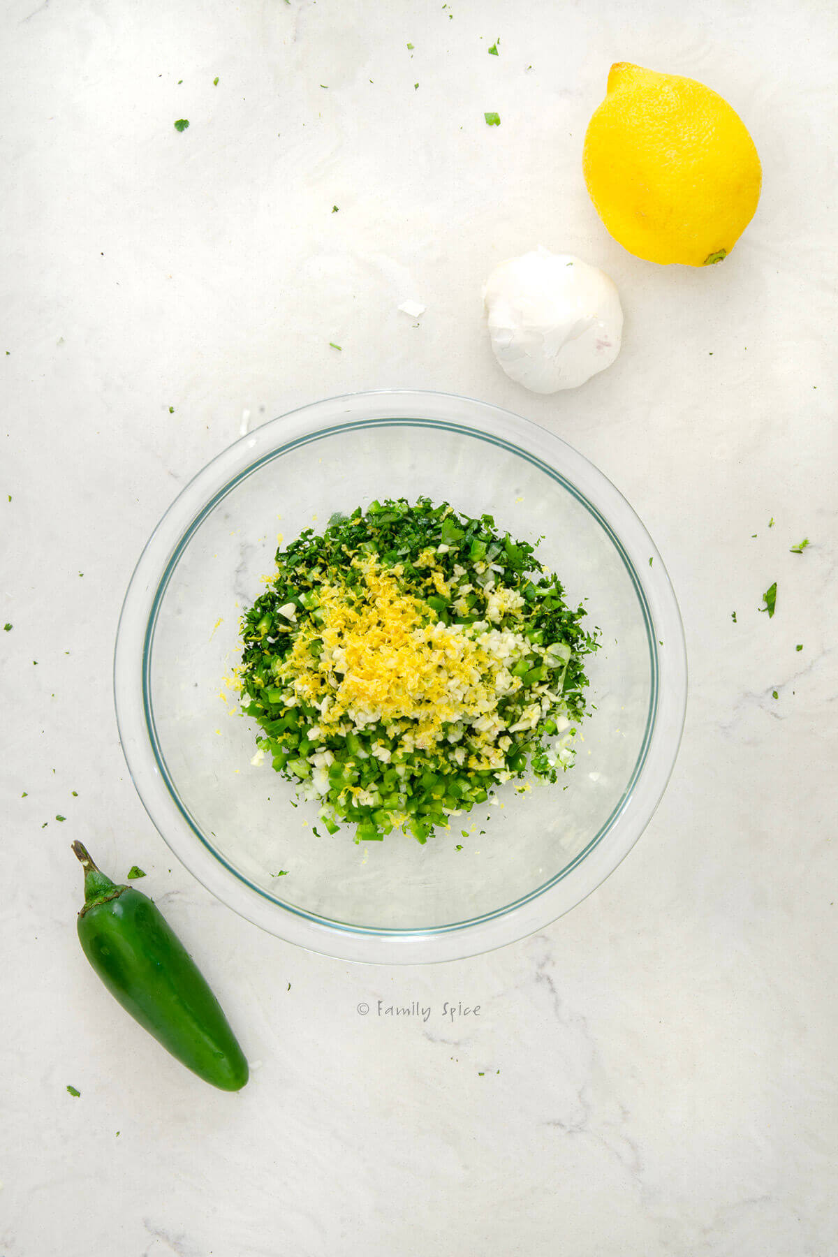 Top view of a small glass bowl with lemon zest added to the rest of the ingredients needed to make cilantro chimichurri with a lemon, jalapeño and garlic next to it