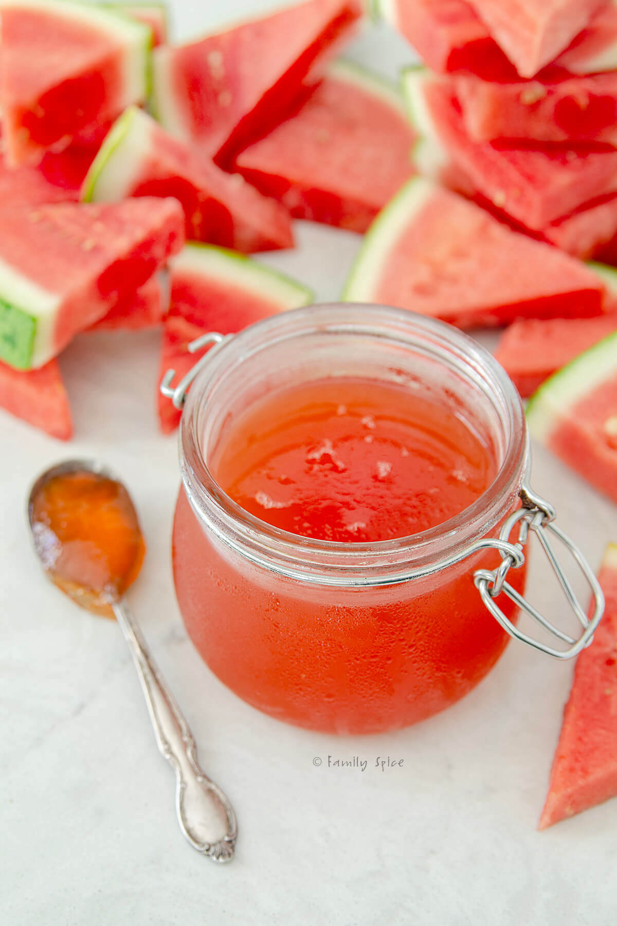 Closeup of a jar of watermelon jelly and a spoon next to it and cut up wedges of watermelon behind it