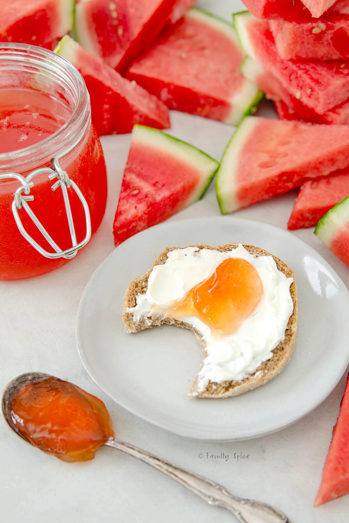 Half a whole wheat English muffin with cream cheese and topped with watermelon jelly with a bite taken out and watermelon behind it