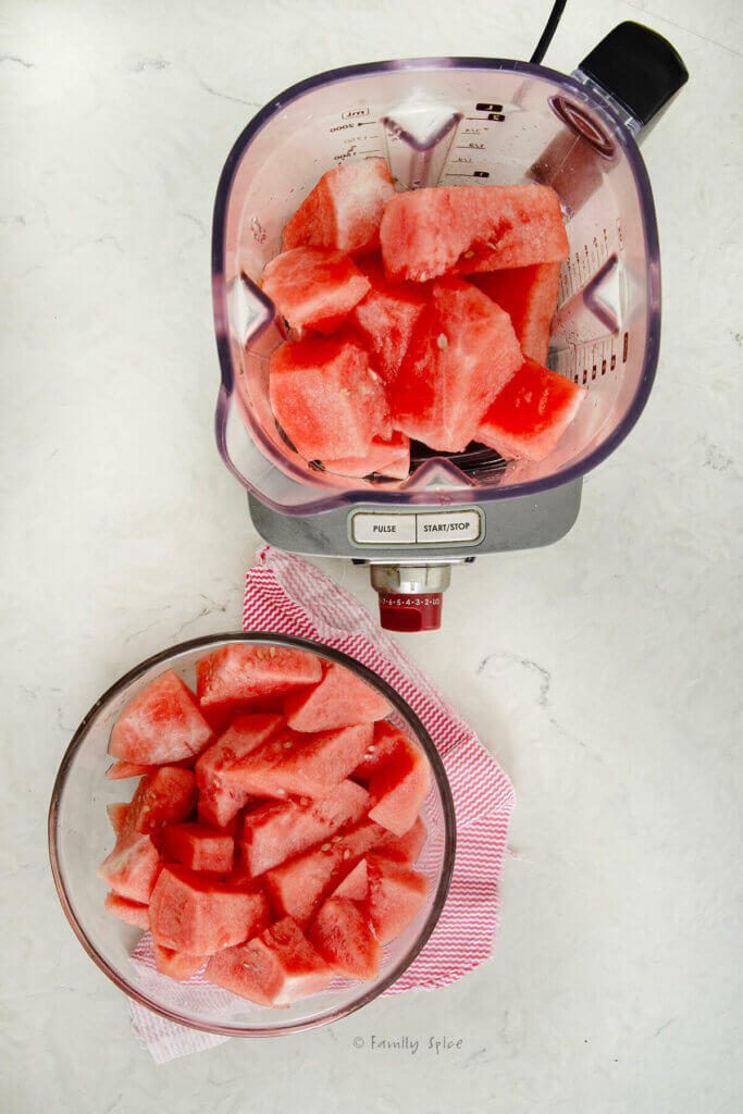 A bowl with cut up pieces of watermelon next to a blender filled with cut up watermelon