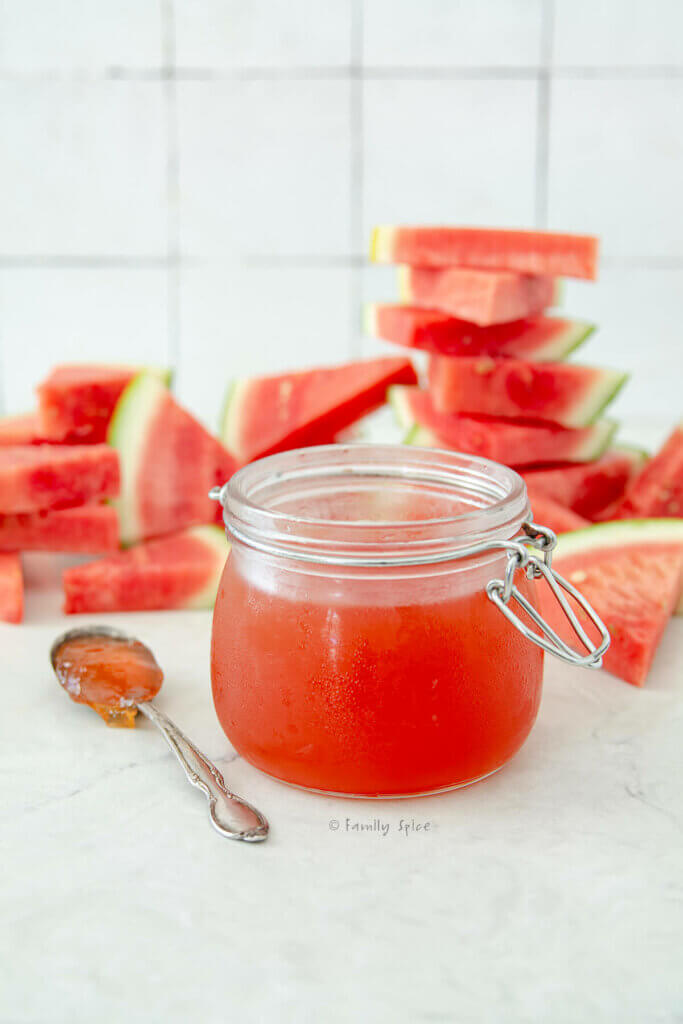 Side view of a jar of watermelon jelly and a spoon next to it and cut up wedges of watermelon behind it