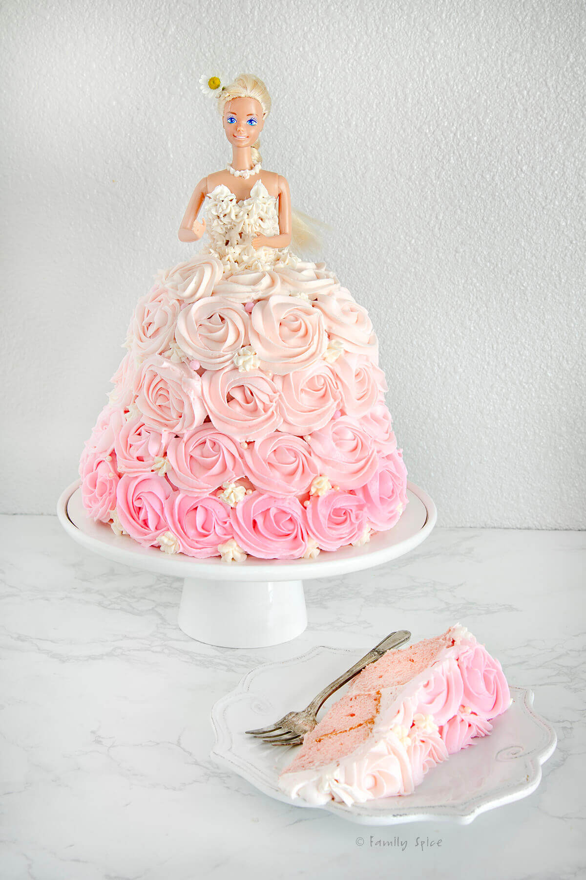 A pink ombre barbie cake on a white cake stand with a slice cut out on a plate