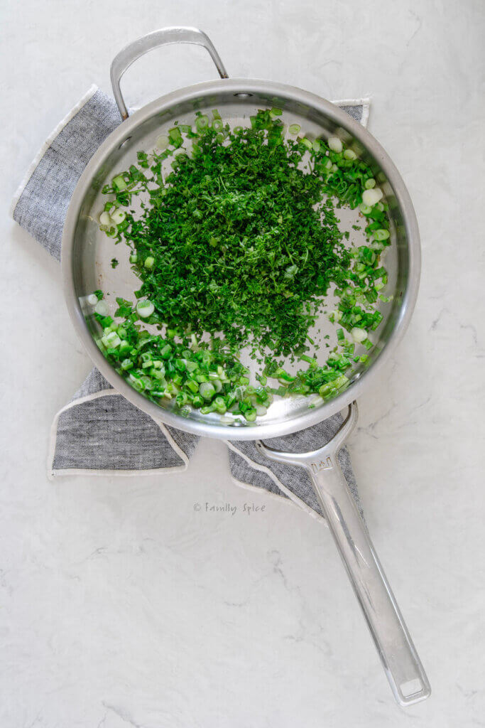 Top view of a stainless pan with chopped green onions and fresh herbs sautéing in it