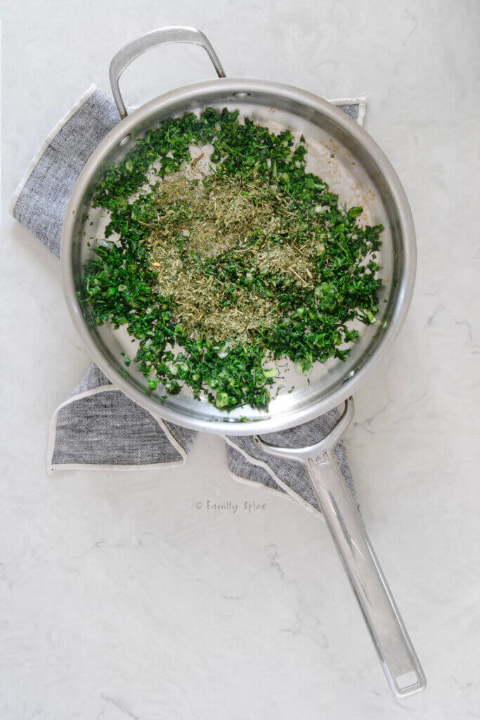 Top view of a stainless pan with assorted fresh herbs sautéing in it and topped with dried fenegreek