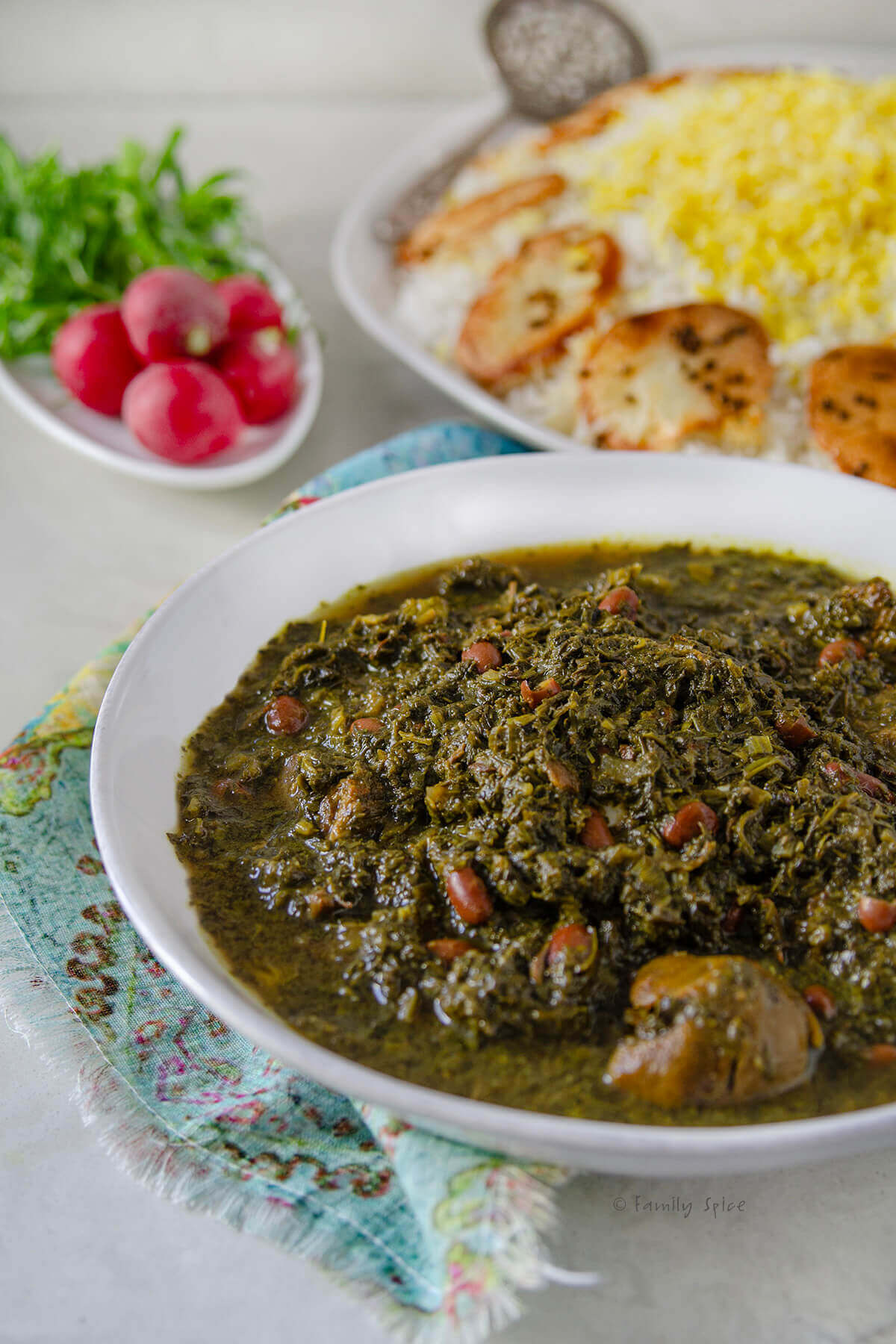 Side view of a bowl of ghormeh sabzi with Persian rice on a platter and fresh radishes and herbs behind it