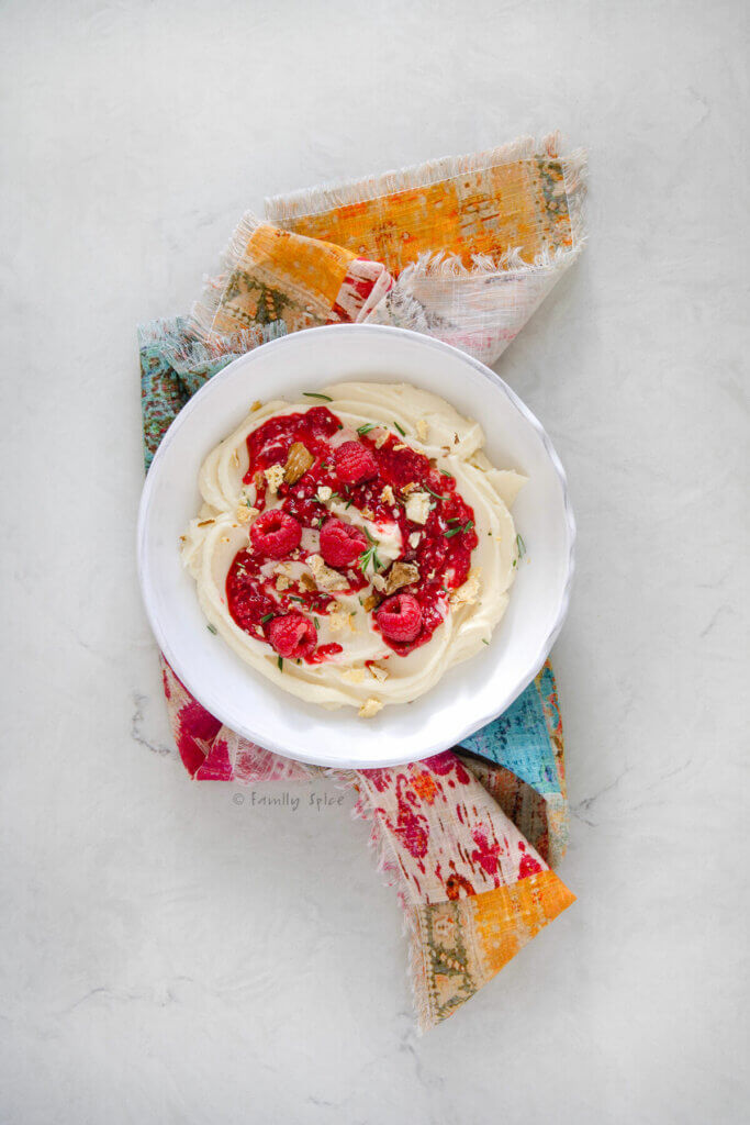Top view of a white shallow bowl with whipped brie in it, topped with raspberry compote, garnished with bits of crunchy brie bits and rosemary on a colorful napkin