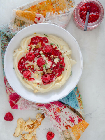 Closeup of a white shallow bowl with whipped brie in it, topped with raspberry compote, garnished with bits of crunchy brie bits and rosemary on a colorful napkin