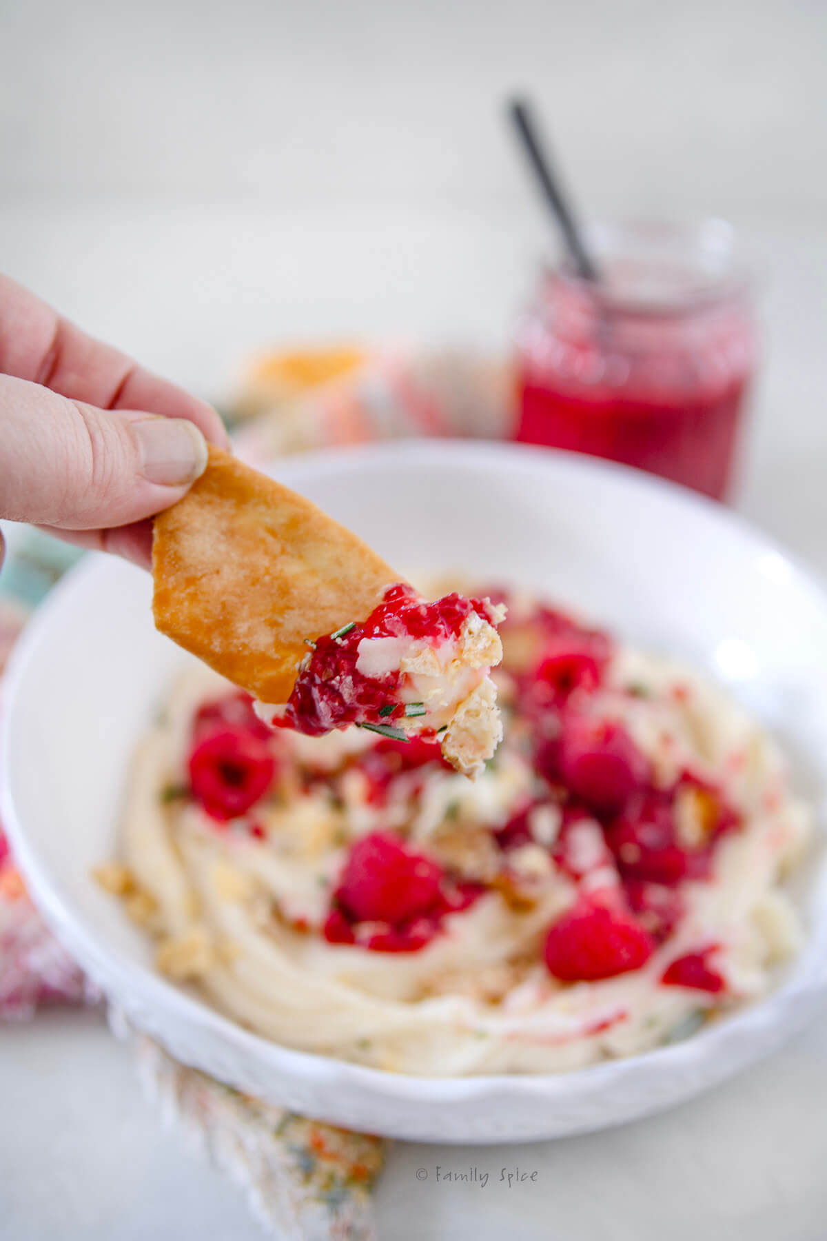Side view of a hand holding a pita whip with whipped brie on it with a white bowl with whipped brie and raspberry compote behind it