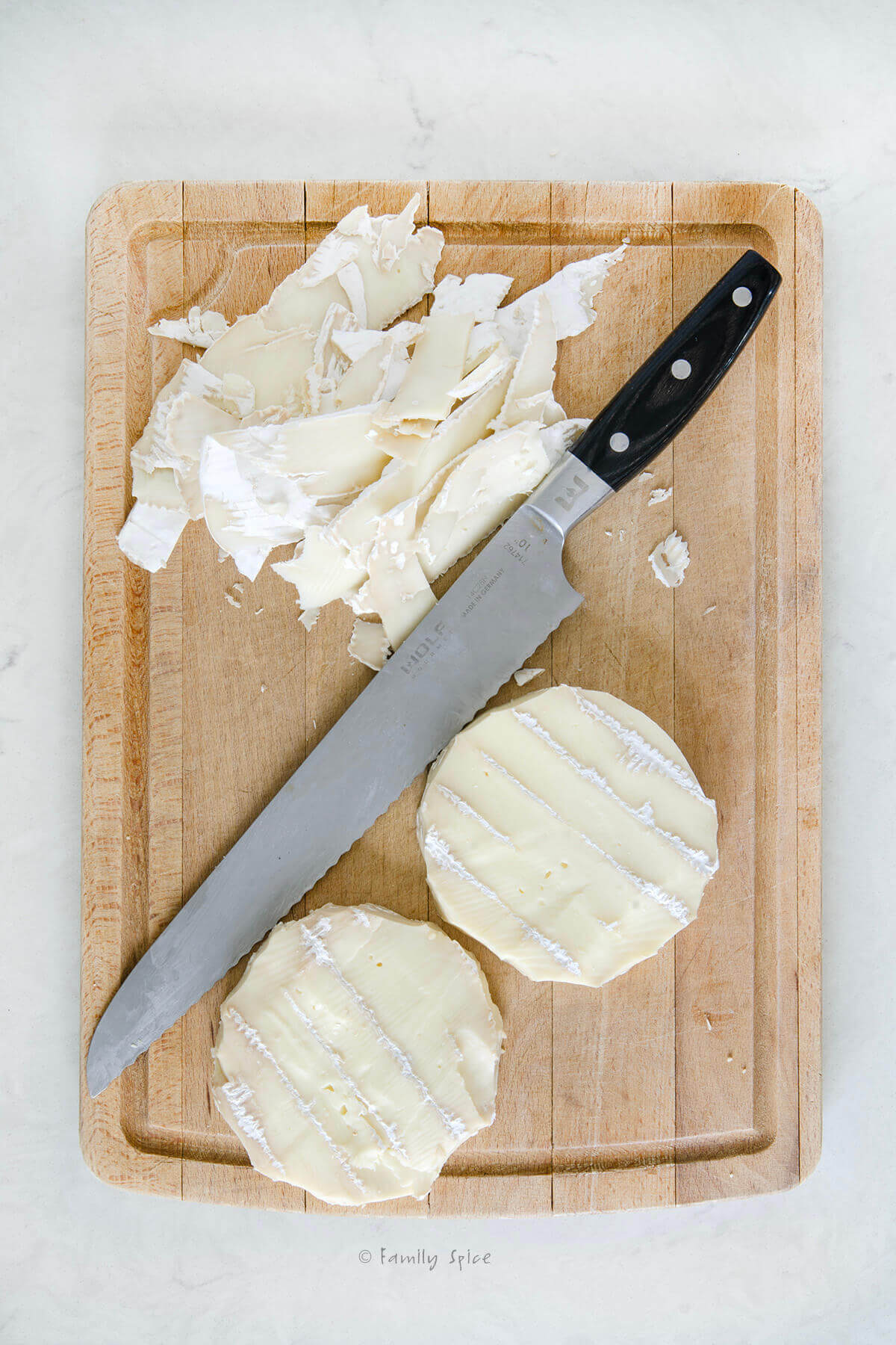 Top view of a cutting board with two wheels of brie with the wax cut off and a serrated knife next to it