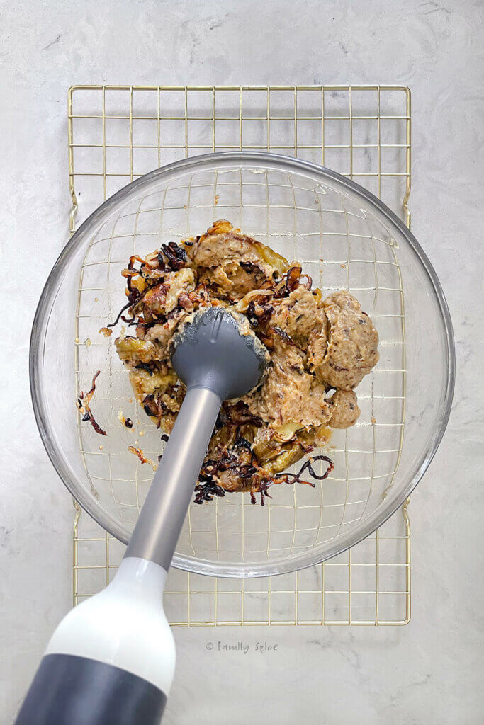 A glass mixing bowl on a cooling rack with an immersion blender combining caramelized onion slice and roasted eggplant slices